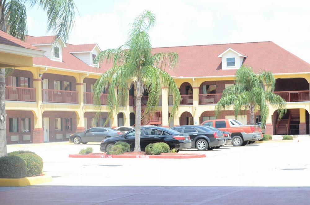 A two-story building with a red roof and yellow walls, featuring a parking lot with several cars and palm trees in the foreground.